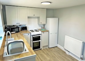 A compact kitchen with white cabinets, a stove, oven, microwave, and a refrigerator. - A beautifully restored two-storey, two bedroom cottage sleeping four people. (Filey)