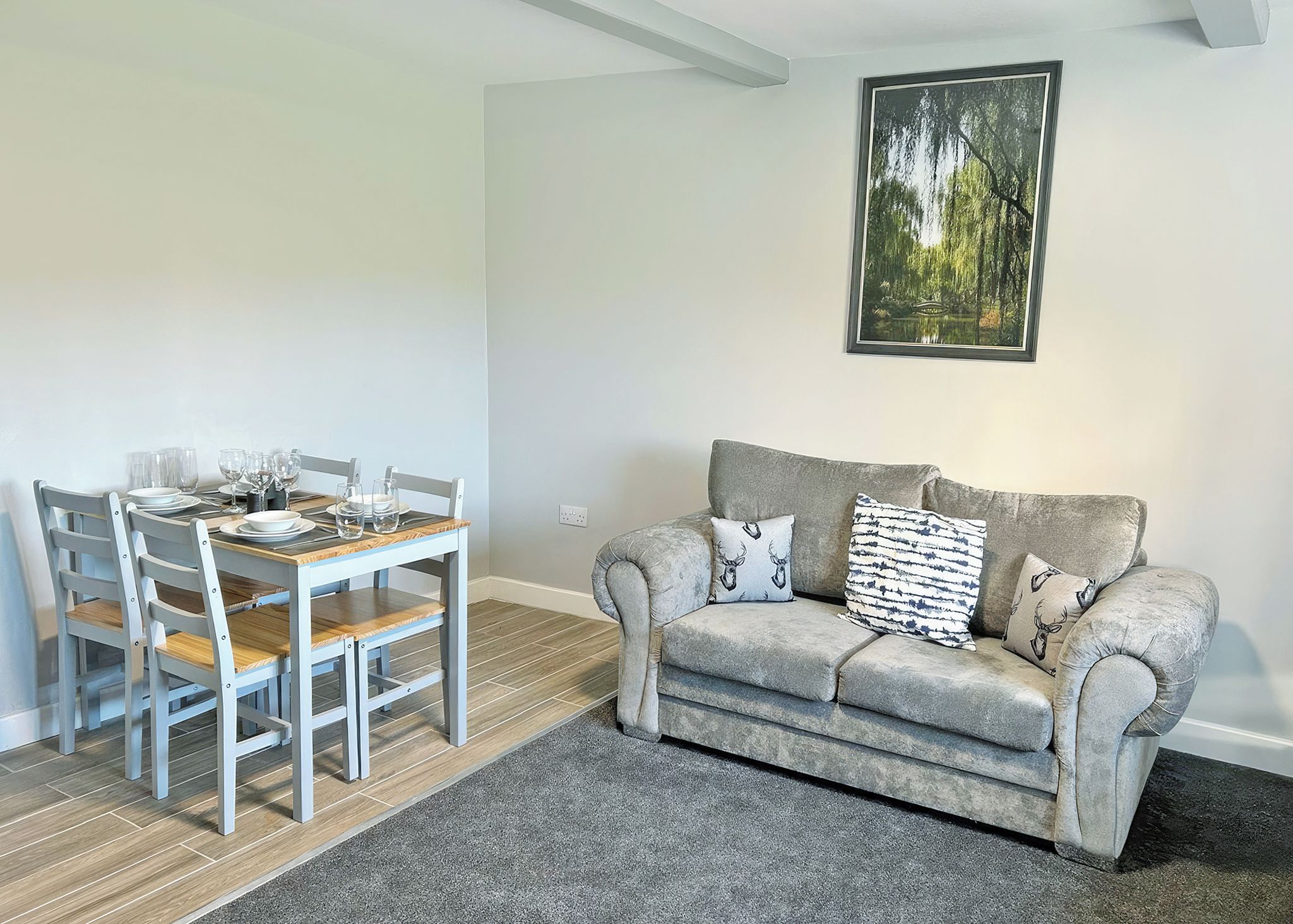 A dining area with a table set for four, a grey sofa with patterned cushions, and a framed landscape painting on the wall.