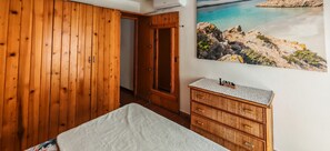 Habitació amb un armari de fusta, un llit amb estriat blanc, un taulell de vimet i una fotografia emmarcada d'un paisatge costaner.