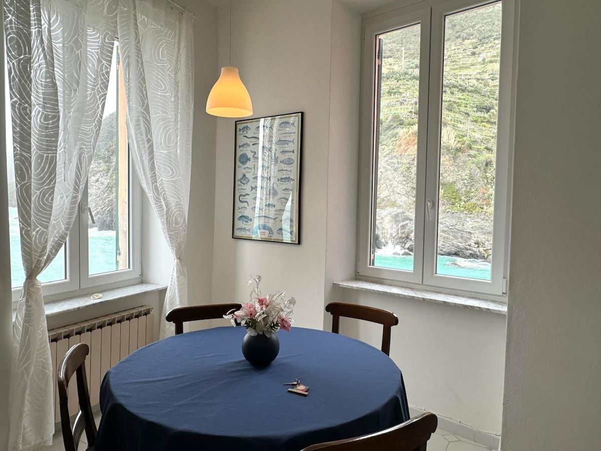 Ein Essbereich mit einem runden Tisch, Stühlen, einer Vase mit Blumen, einem gerahmten Bild an der Wand und Fenstern mit Blick auf Grünflächen.