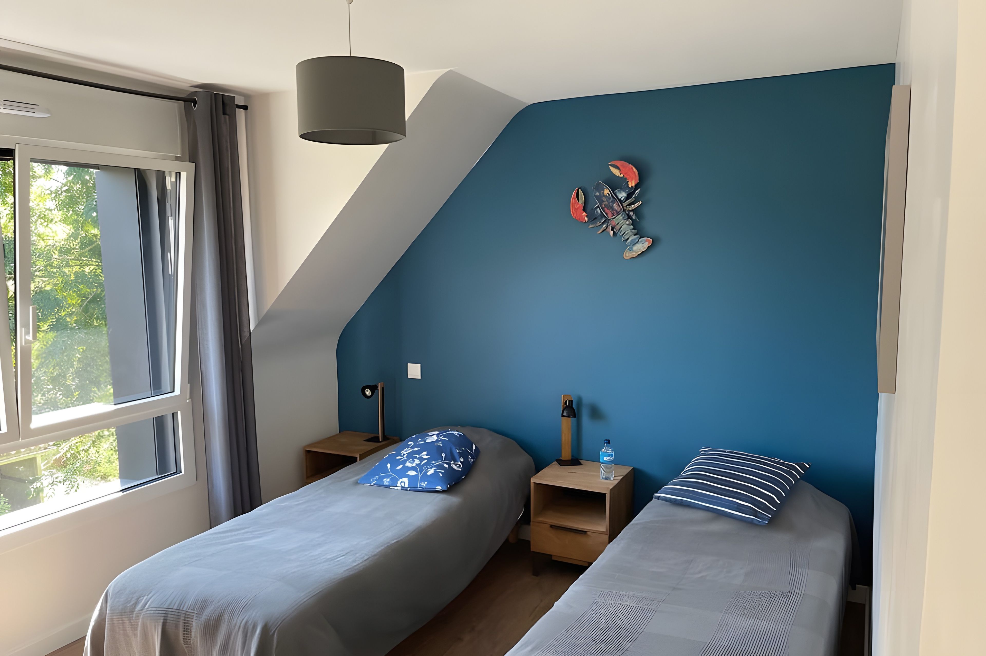 A bedroom with two single beds, a nightstand with a lamp, a wall-mounted shelf, and a window with curtains.