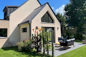 A modern house with a sloped roof, a large window, and a well-maintained garden with various plants and flowers.