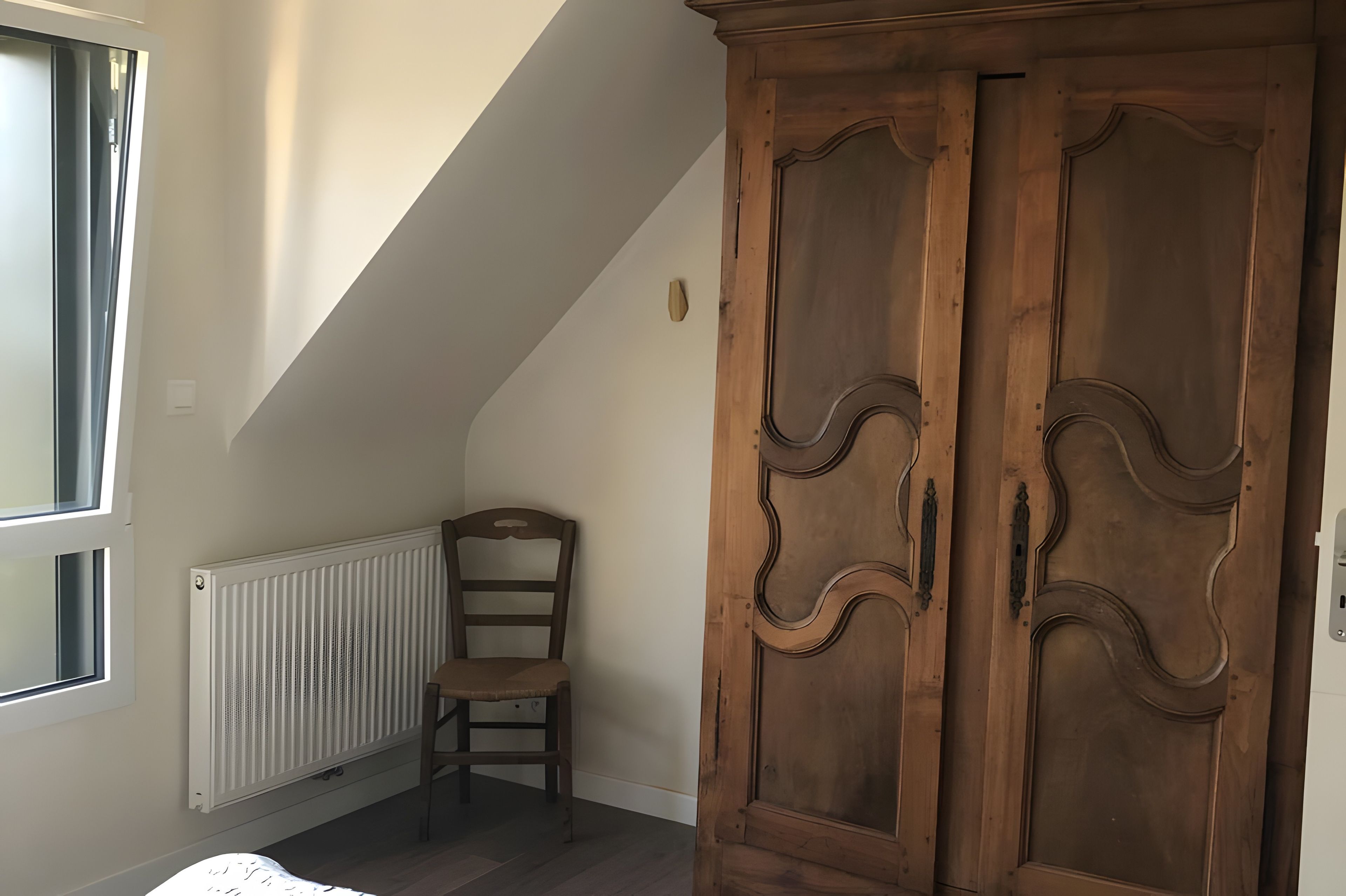 Ein Zimmer mit schrägen Wänden, einem Heizkörper, einem Holzstuhl und einem großen Holzschrank.