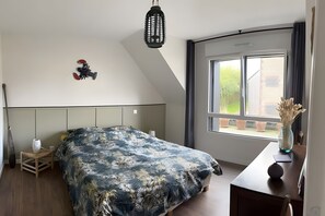 A bedroom with a bed, a hanging light fixture, a window with a view, and a small table with a vase.