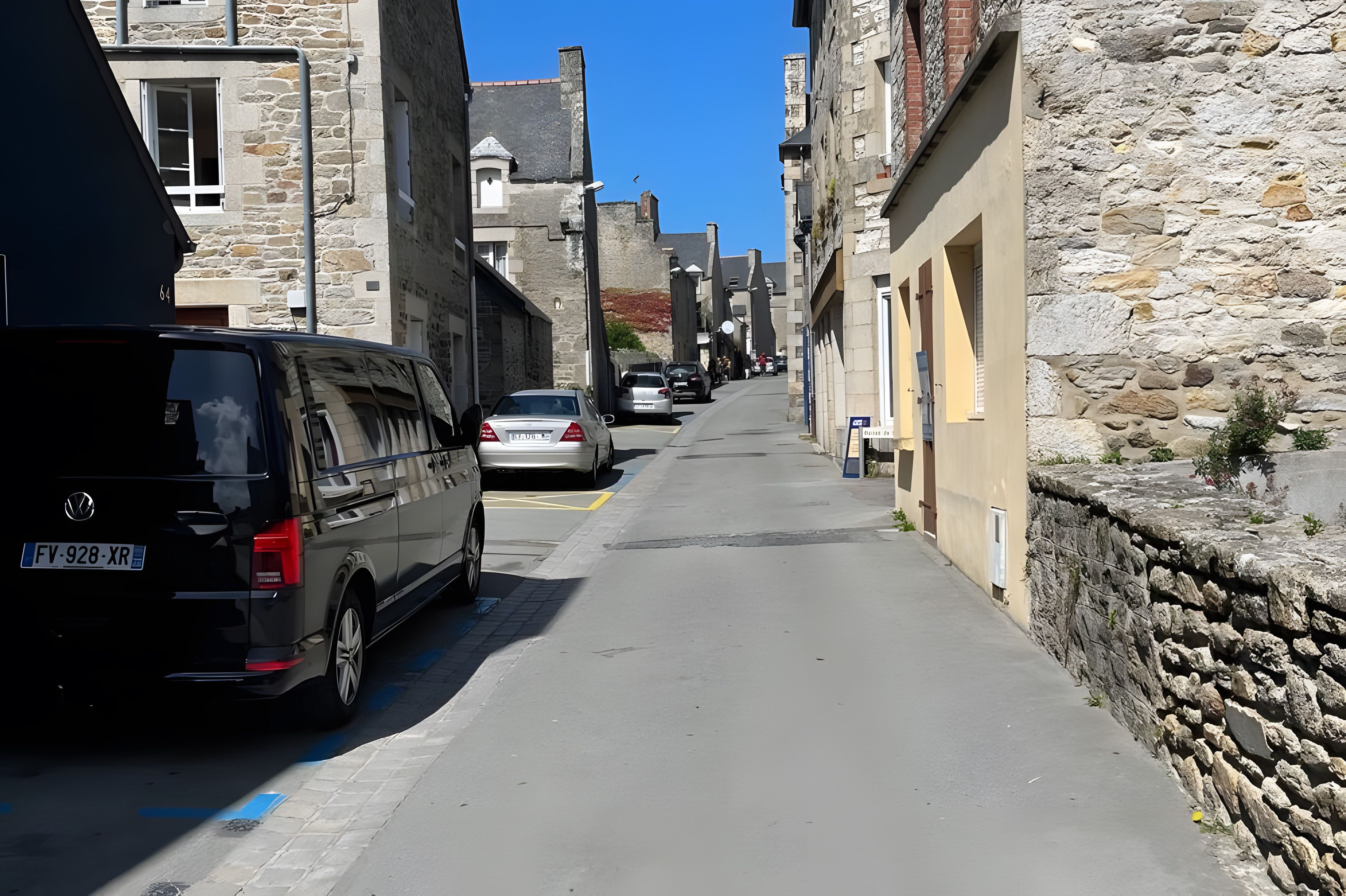 Una calle estrecha con edificios de piedra, coches estacionados y cielo despejado.