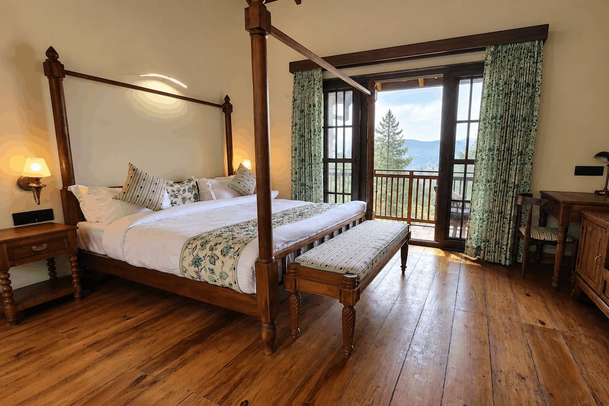 A four-poster bed with a bench, wooden floors, a view through a glass door, and a wooden cabinet.