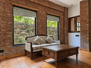 A room with a brick wall, a sofa with floral cushions, a wooden table, and a stone wall outside the window.