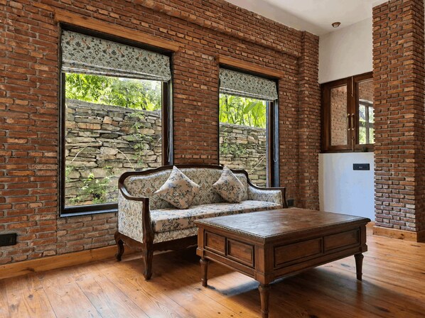 A room with a brick wall, a sofa with floral cushions, a wooden table, and a stone wall outside the window. - The Camellia House Hotel (Almora)