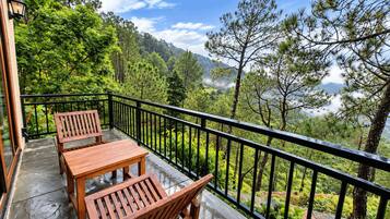 A balcony with a wooden table and chairs, surrounded by a black railing, overlooking a forested landscape.