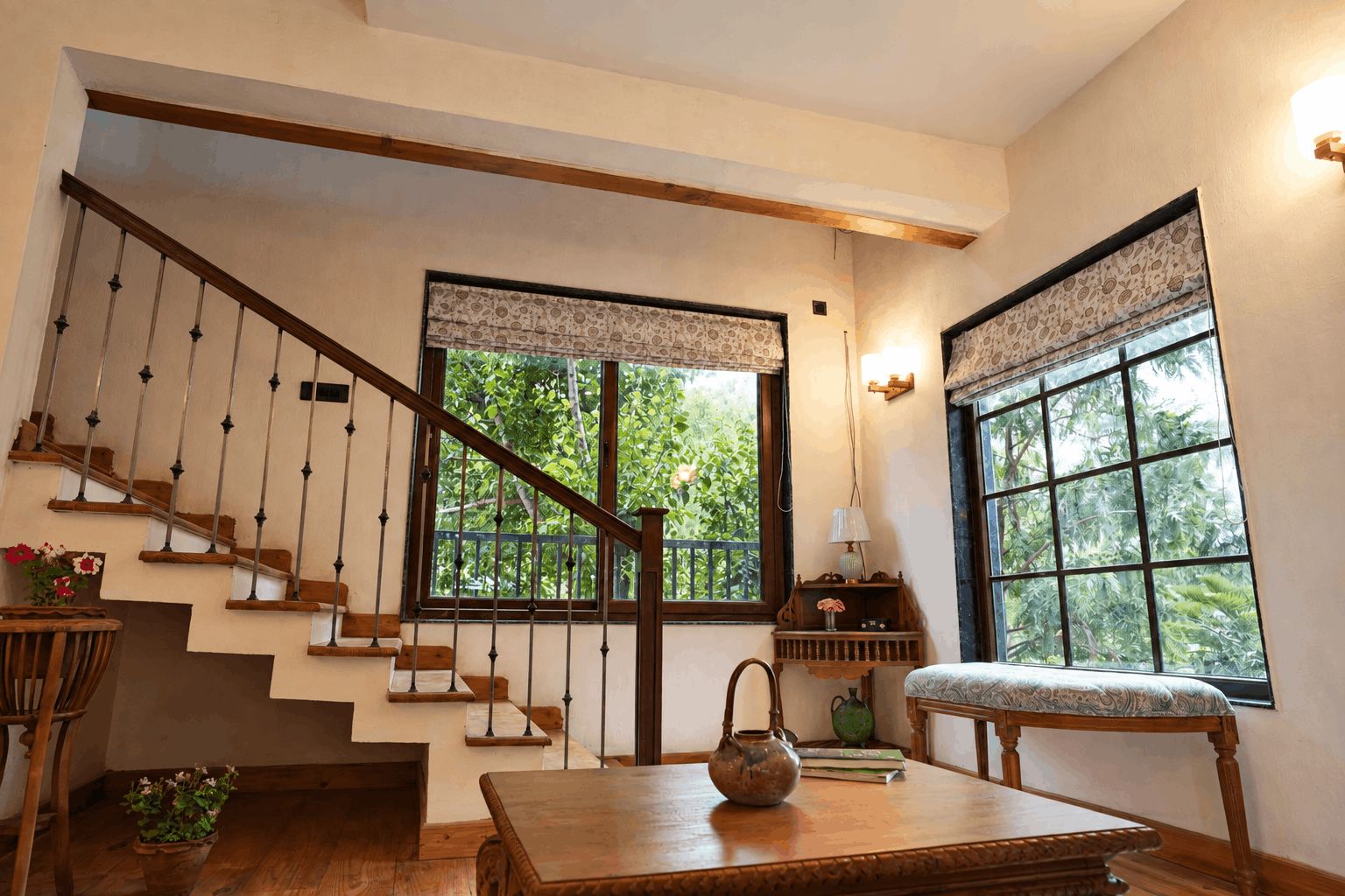 A staircase with wooden steps and metal balusters, a wooden bench, a table with a teapot, and two windows with patterned curtains.