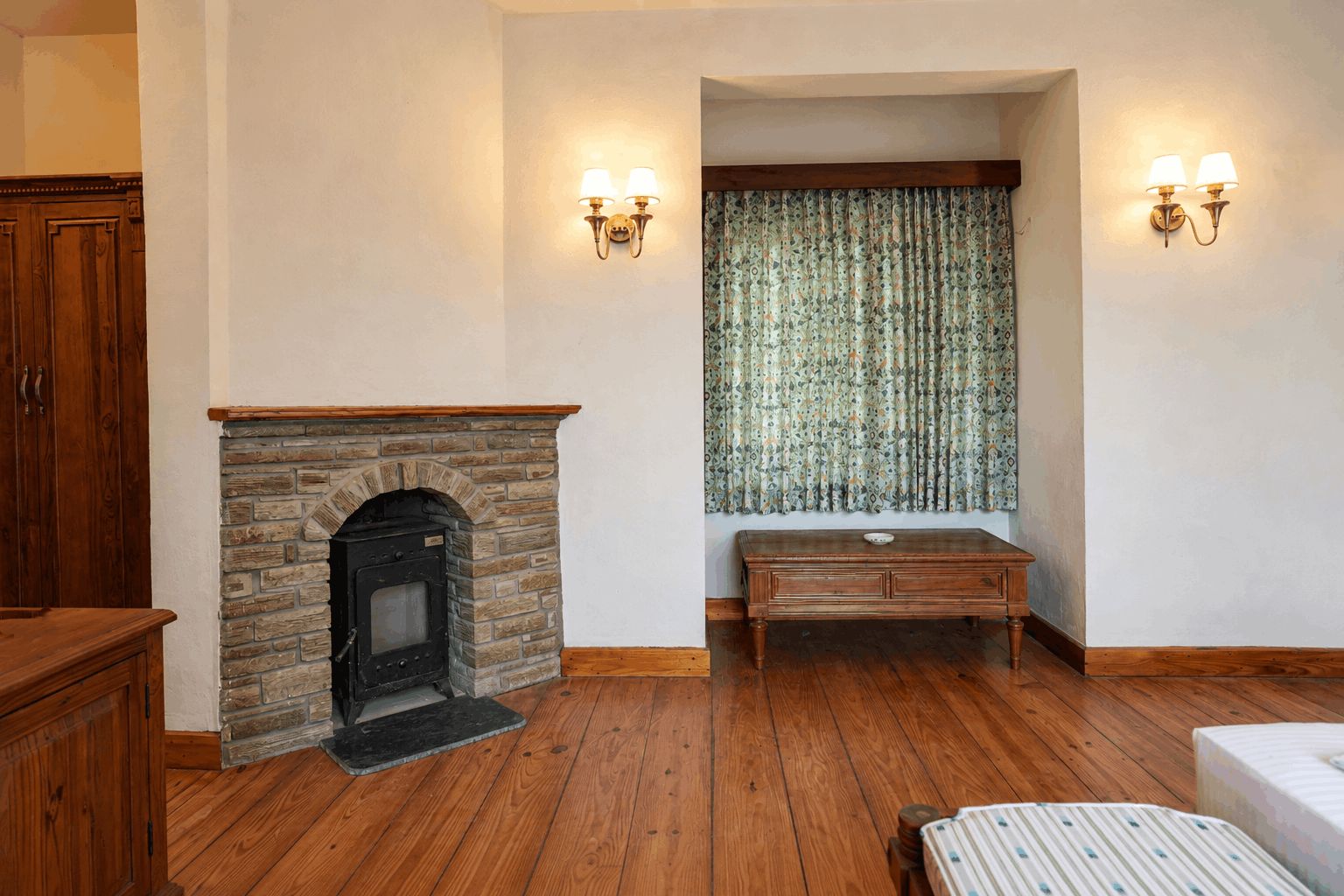 Una habitación con chimenea de piedra, piso de madera, un banco y dos lámparas fijadas a la pared.