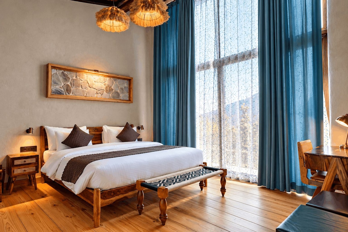 A bedroom with a large bed, wooden headboard, a nightstand, a bedside table, a hanging light fixture, and a view through a window with blue curtains.