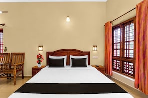 A hotel room with a large bed, wooden headboard, two bedside tables, a nightstand, a chair, a window with curtains, and wall-mounted lights. - Collection O Elephant Camp Thekkady (Thekkady)