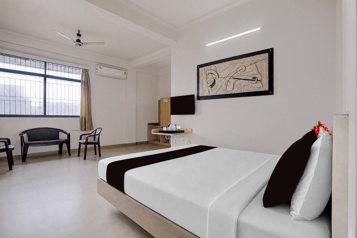 Habitación de hotel con cama, escritorio, silla, ventana con cortinas, unidad de aire acondicionado y una obra de arte colgada en la pared.