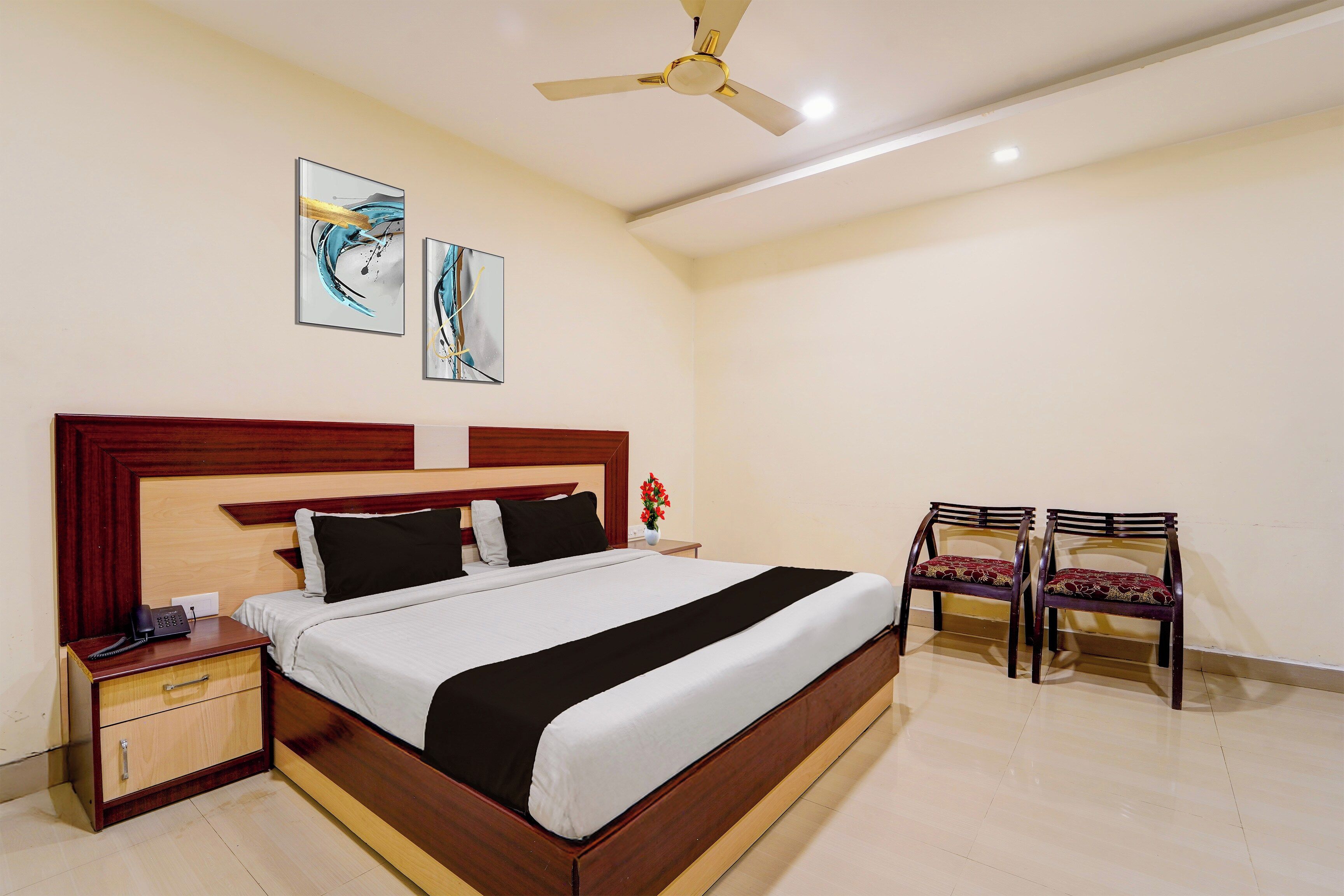 Habitación de hotel con cama, mesitas de noche, escritorio, dos sillas, ventilador de techo y una obra de arte en la pared.