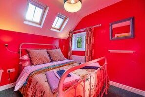 A bedroom with a red wall, a bed with patterned bedding, two skylights, a window with curtains, and a mirror on the wall. - Finest Retreats | Ffrith-Bach (Abergele)