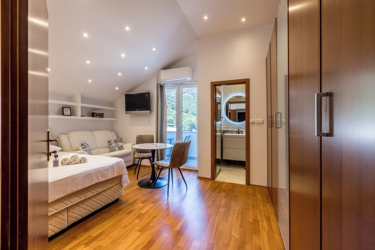 A modern bedroom with a bed, a TV, a dining area, and a view of the outdoors.