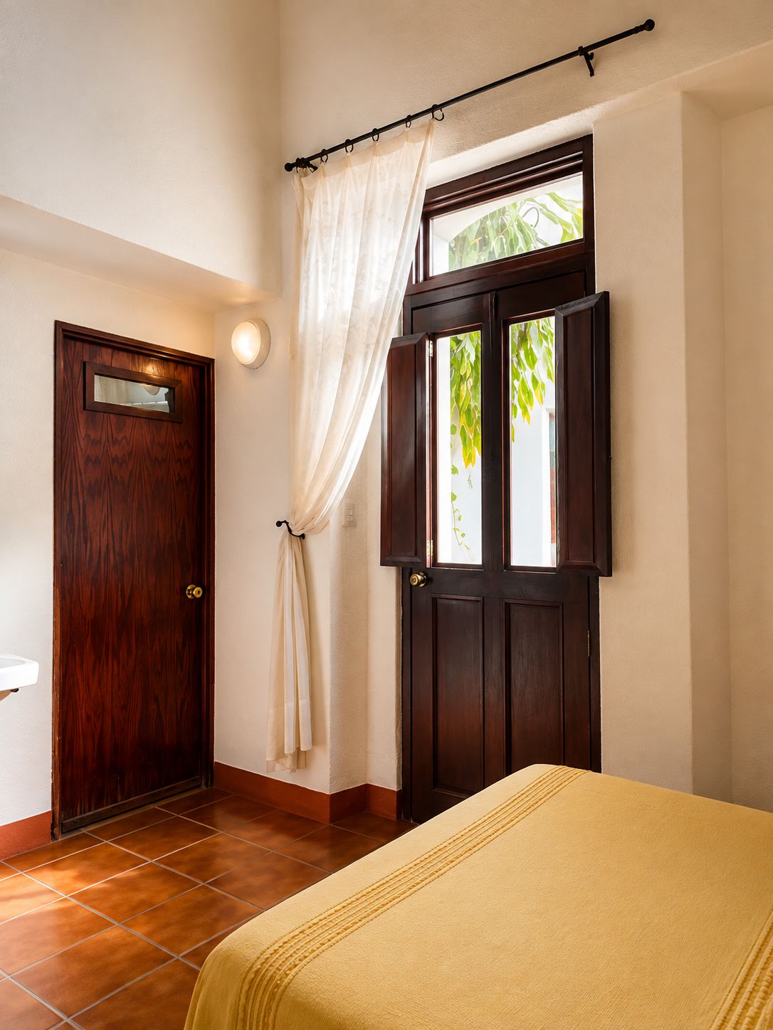 A room with a wooden door, a window with a view of greenery, and a bed with a yellow bedspread.