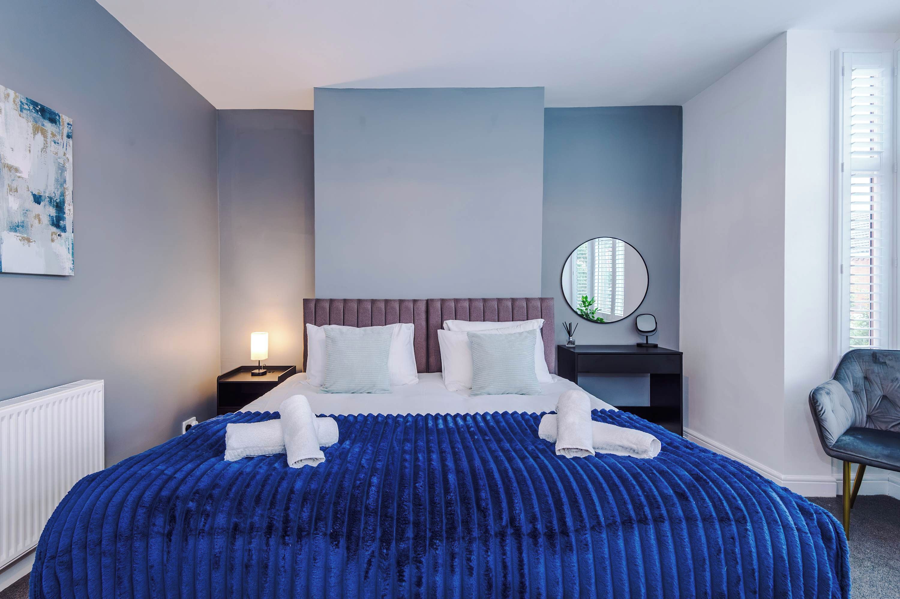 A hotel room with a bed, a nightstand, a chair, a round mirror, and a painting on the wall.