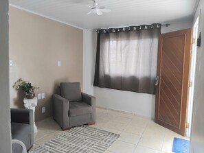 Ein Zimmer mit grauem Sessel, einem gemusterten Teppich, einem Fenster mit Vorhang und einer Holztür.