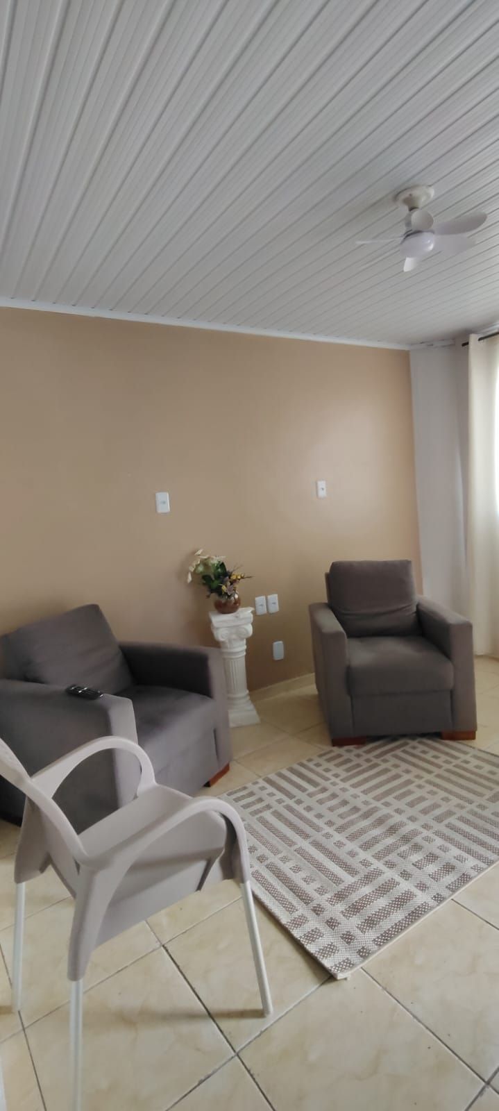 A living room with a beige wall, a grey sofa, a grey armchair, a white plastic chair, a patterned rug, and a ceiling fan.