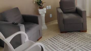 A living room with a beige wall, a grey sofa, a grey armchair, a white plastic chair, a patterned rug, and a ceiling fan.
