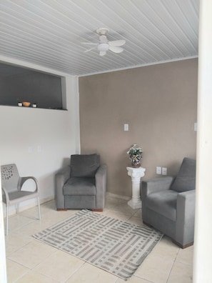 Una habitación con ventilador de techo, dos sillones grises, una mesita blanca con un florero y una alfombra estampada.
