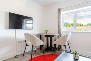 Ein modernes Zimmer mit flachbildfernseher an der Wand, zwei Polsterstühle, einem kleinen Tisch mit Topfpflanze und einem Fenster mit Blick auf Grünflächen.