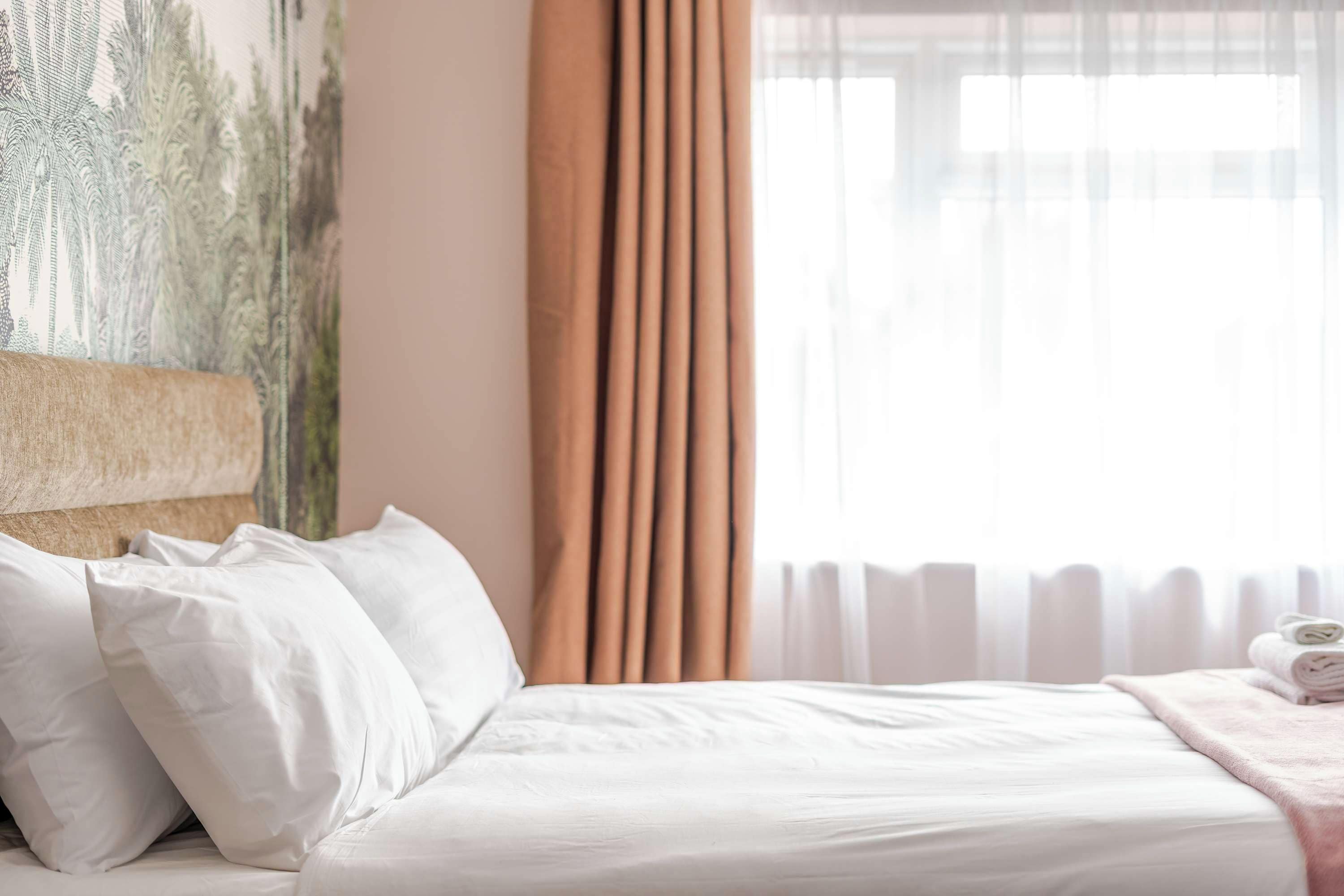 A neatly made bed with white linens, a headboard with a textured design, and a window with sheer curtains.