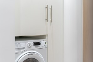 A compact, white washing machine with a digital display and control panel, housed in a minimalist cabinet with a wooden finish. - Stylish 1 bedroom flat in Crawley (London)
