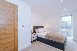 A bedroom with a wooden door, a bed with a headboard, a nightstand, a lamp, and a window with blinds. - 1BR | Free Wi-Fi | Central Stay | Richmond Hill (Leeds)
