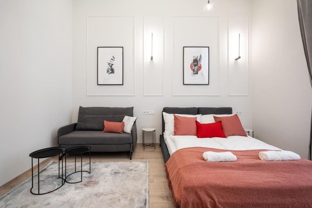 Quarto moderno com sofá cinza, cama com roupa de cama vermelha e branca, e duas imagens emolduradas na parede.