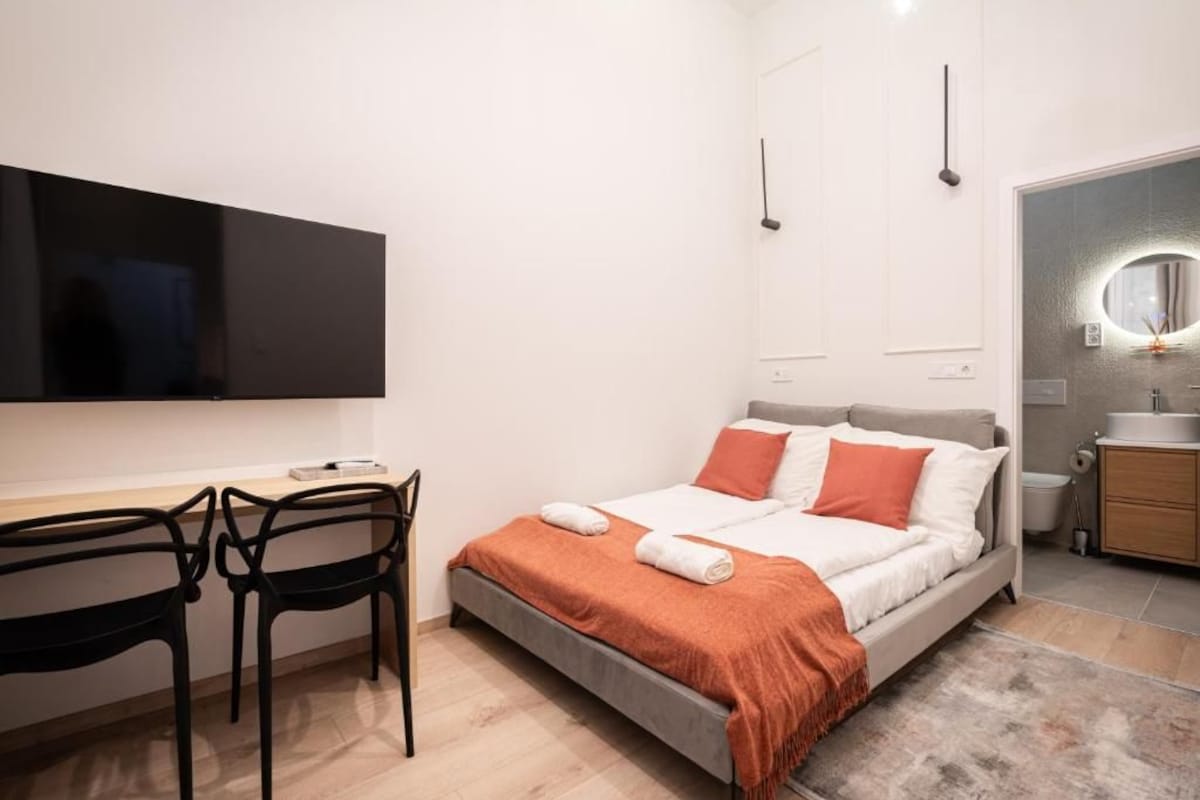 Ein modernes Schlafzimmer mit Bett, Schreibtisch, zwei Stühlen, Fernseher und einem Badezimmer mit Spiegel.