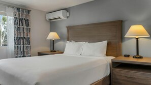 Una habitación de hotel con una cama grande, dos mesitas de noche con lámparas, un aire acondicionado instalado en la pared y una ventana con cortinas.