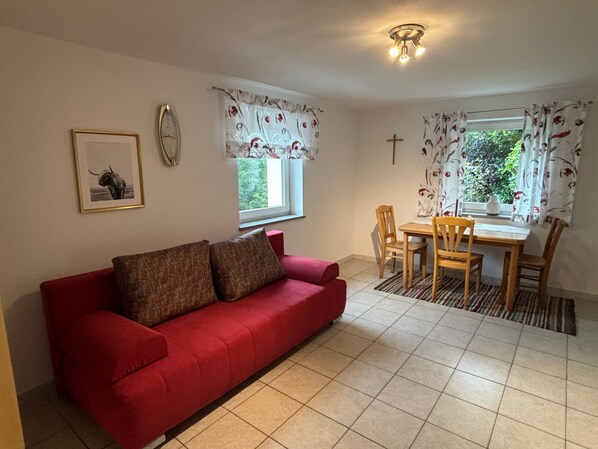 A red sofa with two cushions, a dining table with four chairs, a window with curtains, a wall clock, and a framed picture. - Apartment 'Klein' with Shared Terrace and Shared Garden (Waldkirchen)