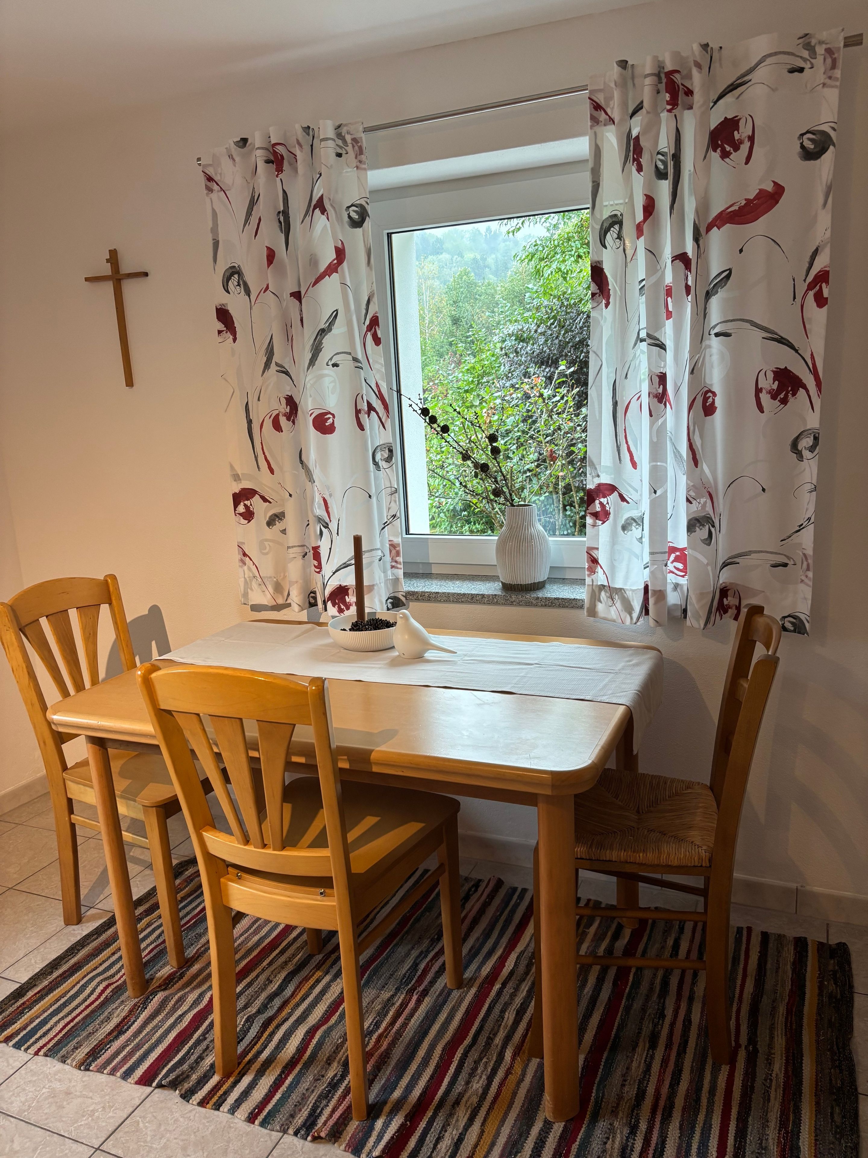 A dining area with a table and chairs, a window with curtains, a vase with flowers, and a cross on the wall.