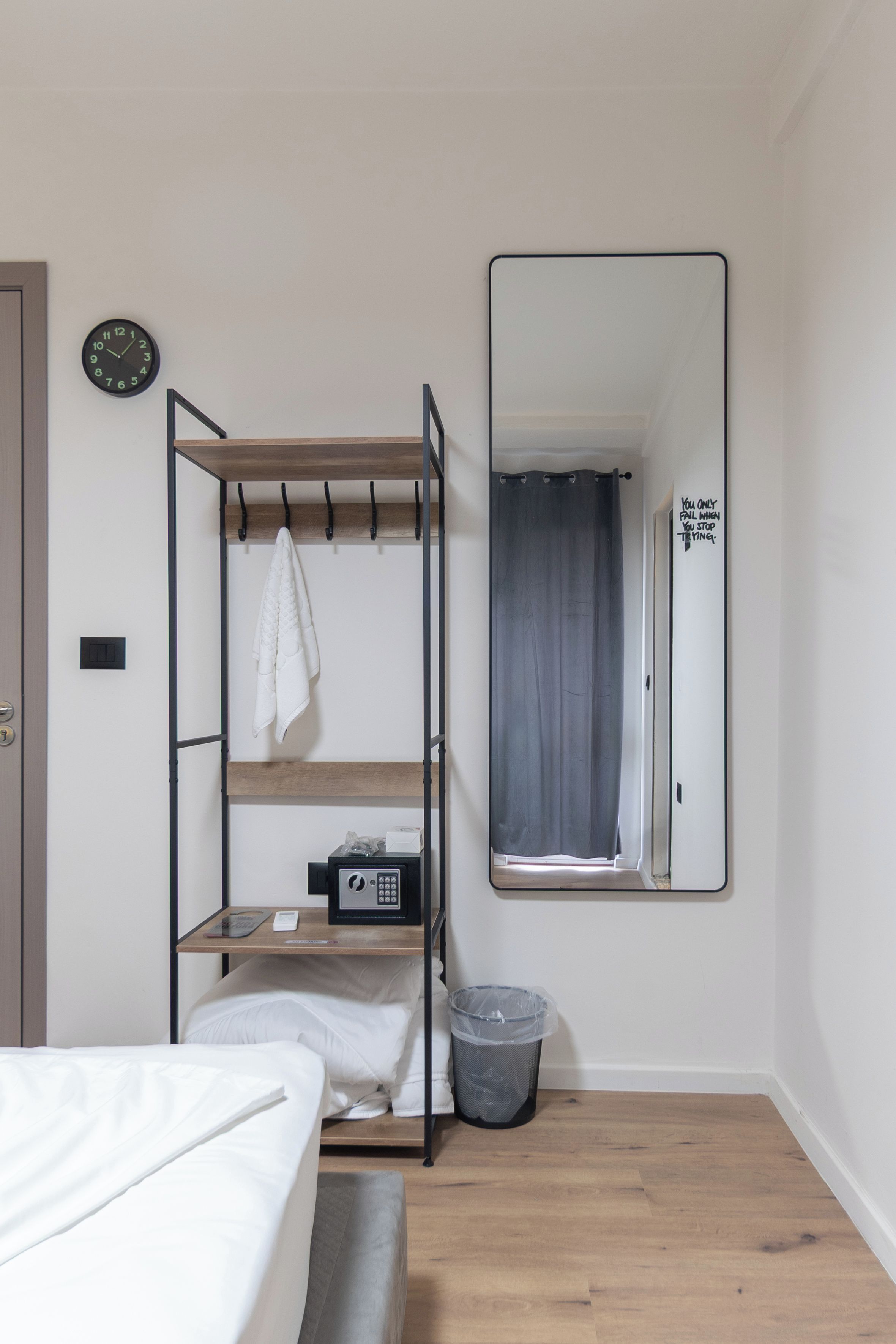 Habitación de hotel compacta con una litera, una mesita de noche con lámpara, un espejo, un cubo de basura pequeño y una puerta con una frase motivacional.