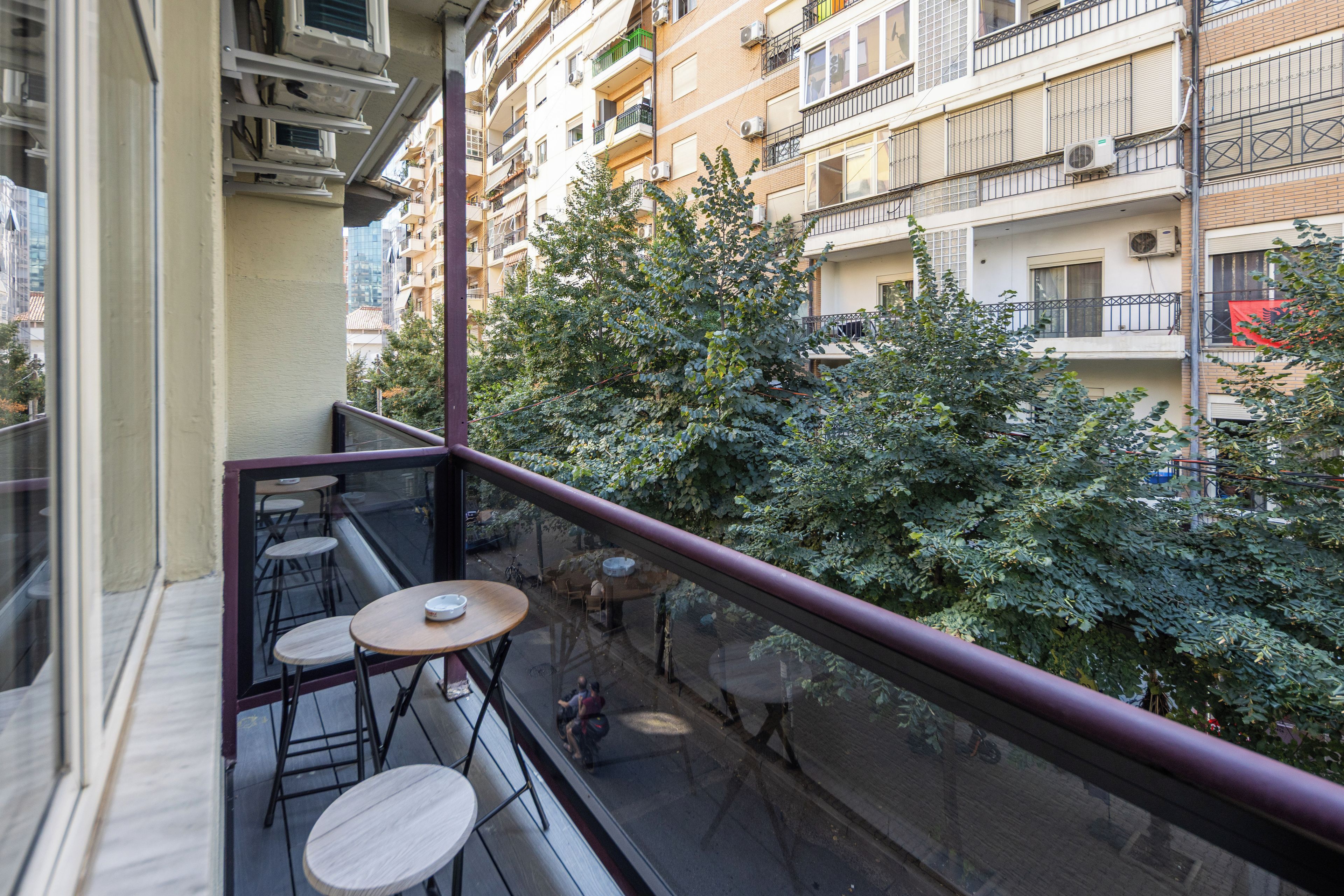 Un balcón con una mesa y sillas, con vista a una calle con árboles y edificios.