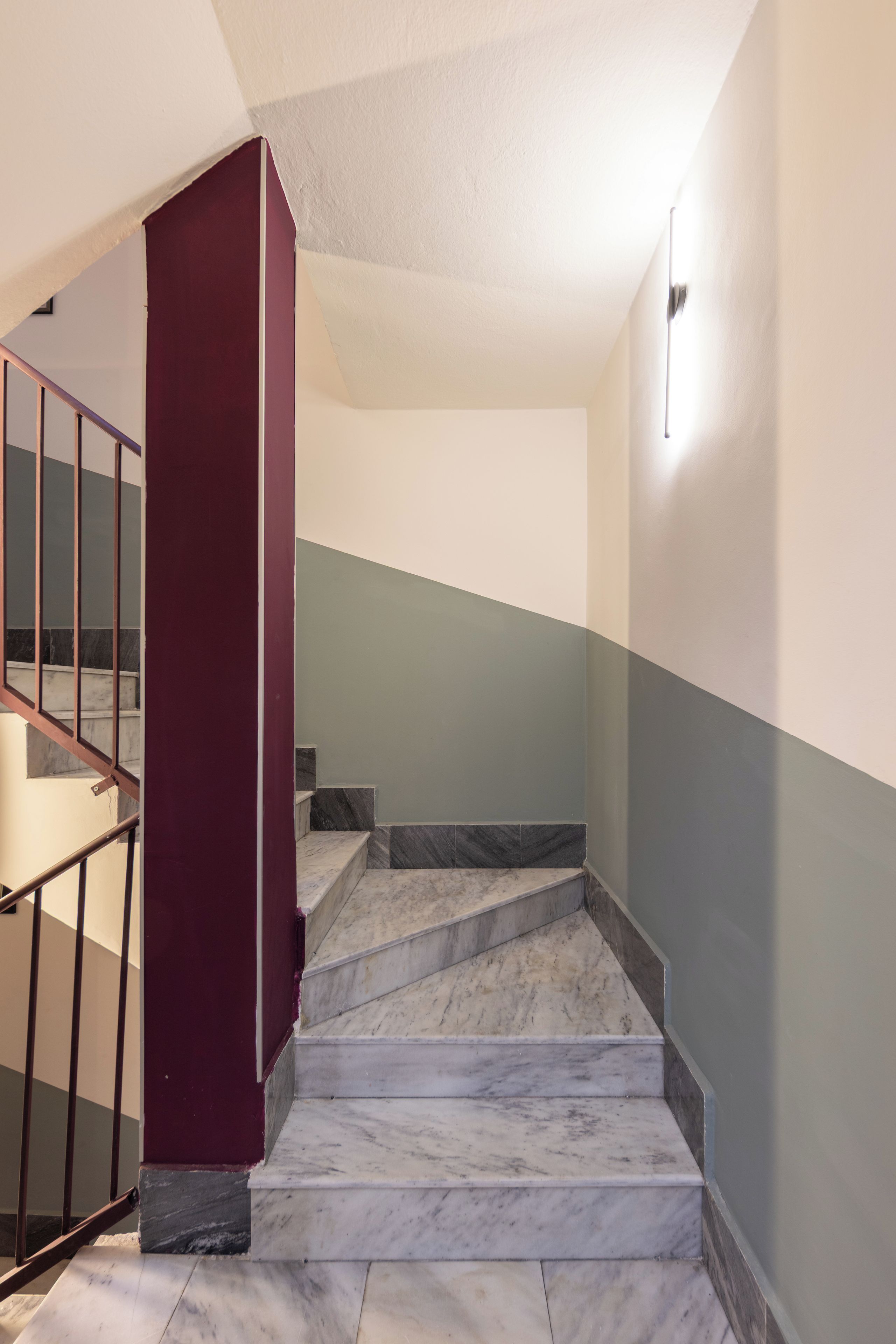Una escalera con peldaños de mármol y barandilla metálica, que conduce a un rellano con una pared de acento rojo.