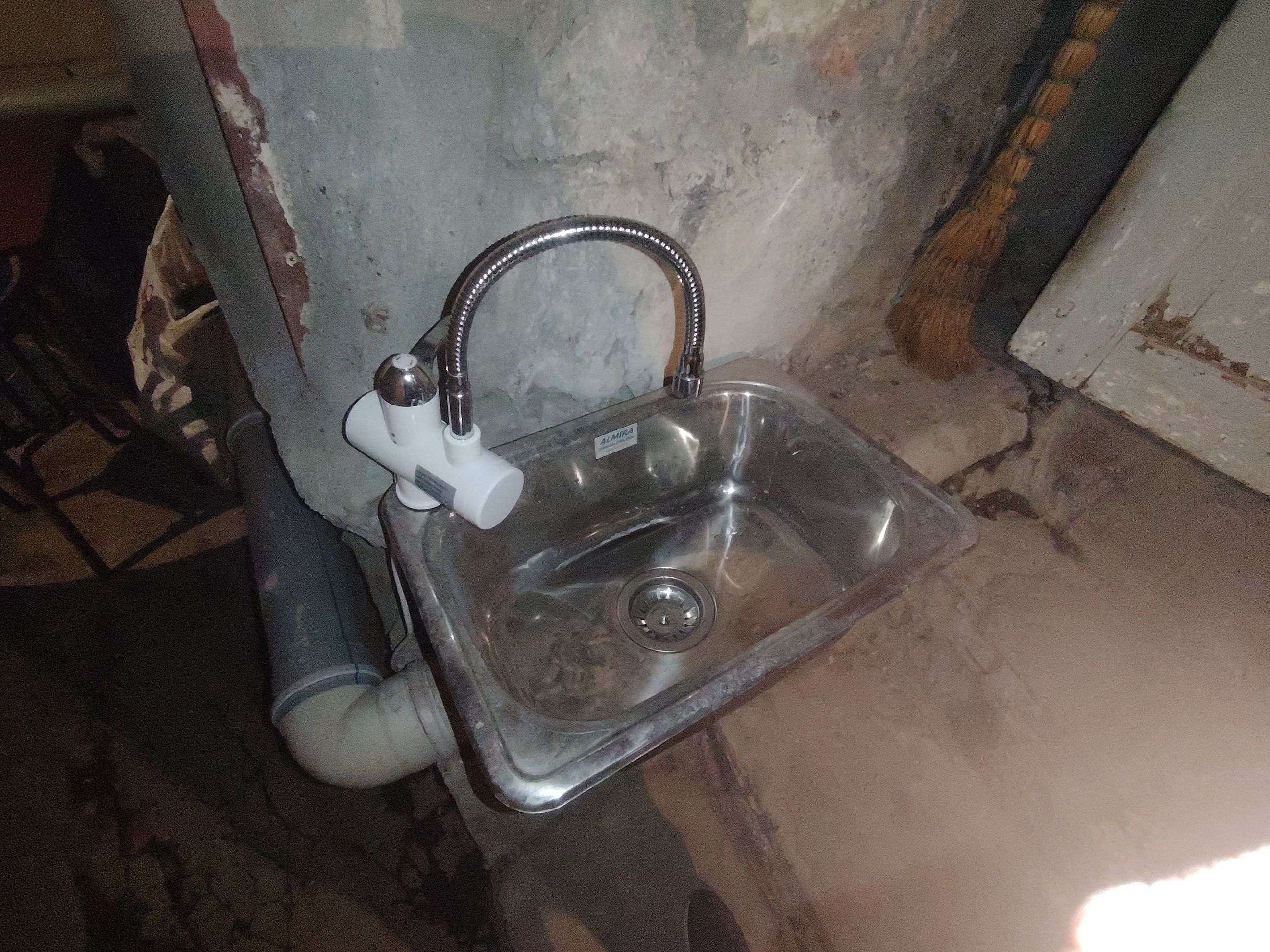 A metallic sink with a faucet and a pipe, mounted on a wall with visible wear and discoloration.