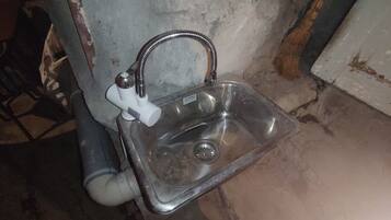 A metallic sink with a faucet and a pipe, mounted on a wall with visible wear and discoloration.
