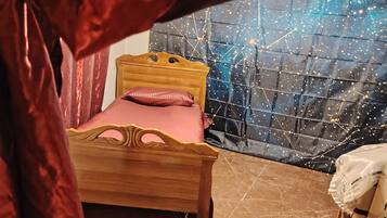A wooden bed with a pink pillow and a wooden headboard, a nightstand with a lamp, and a wall with a starry pattern.