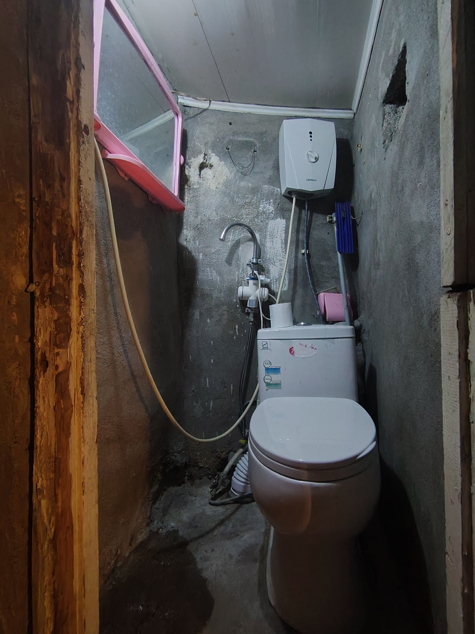Une petite salle de bain peu éclairée, équipée de toilettes, d’un chauffe-eau et d’une fenêtre.