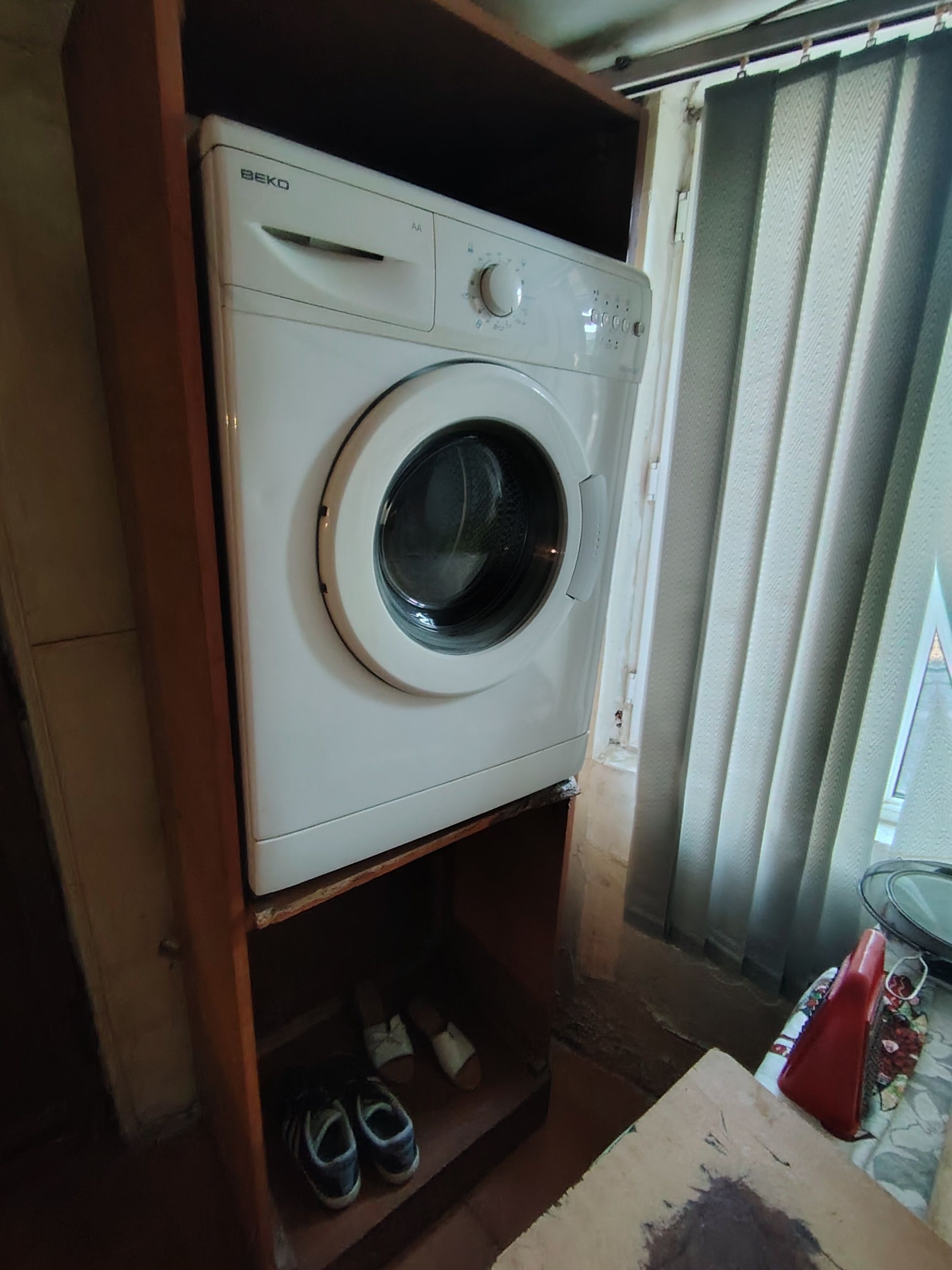 Een witte BEKO wasmachine in een wasruimte met een houten plank eronder.
