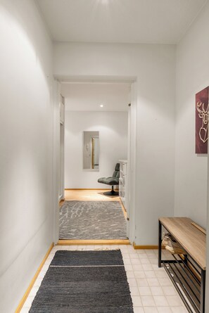A hallway with a grey rug, a bench, and a door leading to a room with a chair and a cabinet. - Arctic Riverside Retreat – Private Sauna - Central (Rovaniemi)
