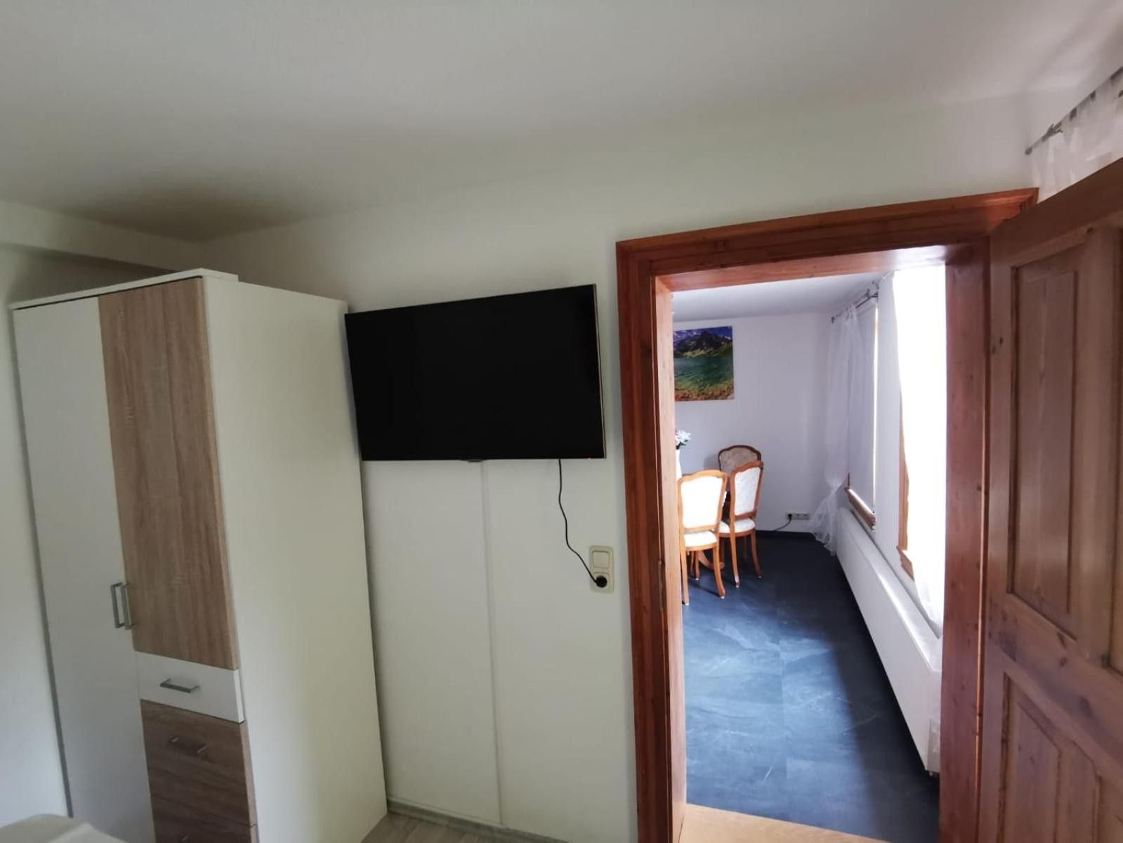A room with a wardrobe, a flat-screen TV mounted on the wall, a dining table with chairs, and a painting on the wall.
