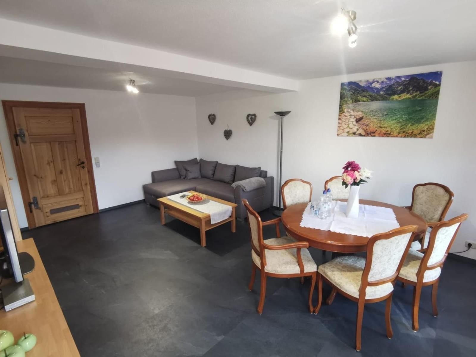 A living room with a sofa, coffee table, dining table with chairs, a painting on the wall, and a door.