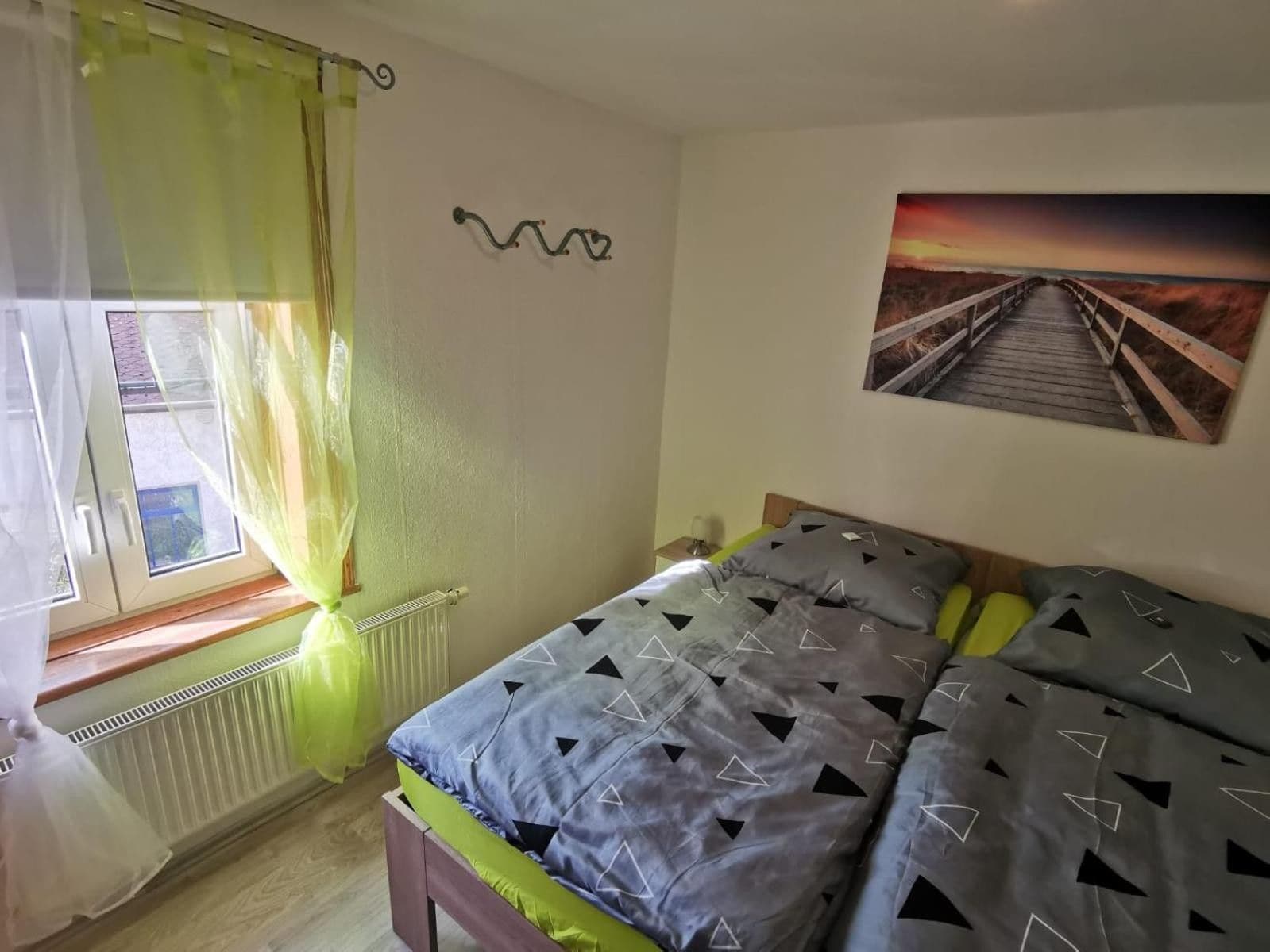 Ein Schlafzimmer mit Bett, einem Fenster mit Vorhängen, einem Heizkörper, einem Wandbild und einem Nachttisch mit Lampe.