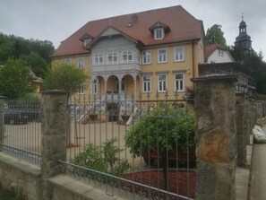 A two-story building with a red-tiled roof, a balcony, and a stone fence.