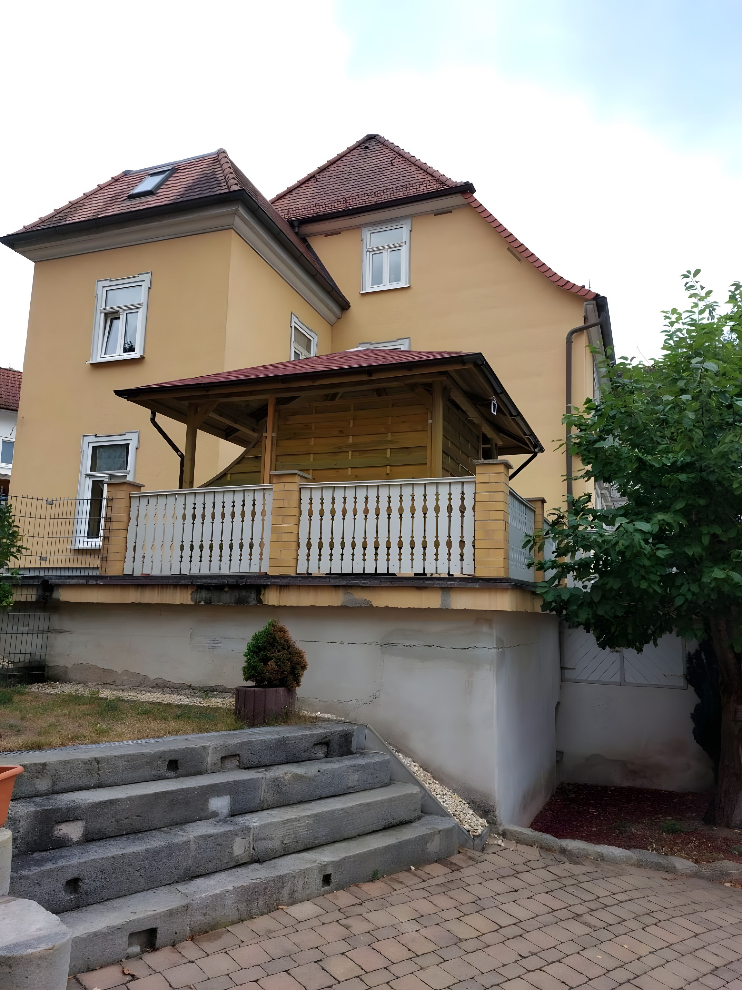 Ein zweistöckiges Gebäude mit einem Balkon, einem kleinen Garten und einer Treppe aus Beton, die zum Eingang führt.
